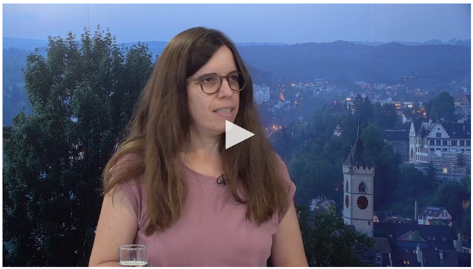 Frau mit langen, glatten braunen Haaren und Brille. Trägt ein Rosa T-Schirt. In Interviewsituation vor Stadtbild als Hintergrund.